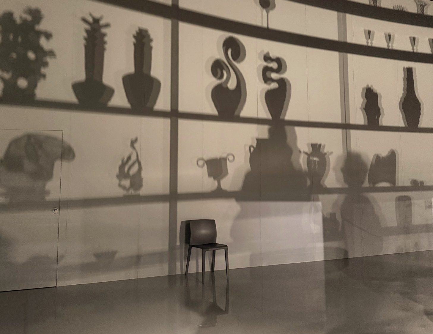 Shadows of vessels and objects on a wall, with a single chair facing them